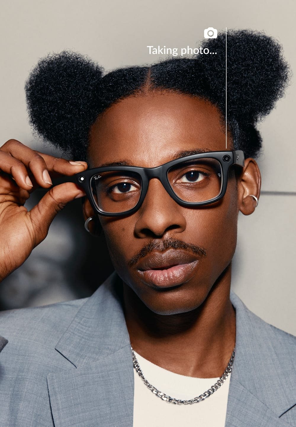 Lunettes rectangulaires noires avec des montures épaisses, verres transparents et un logo à l'avant. Le porteur a des cheveux texturés et porte un costume gris clair.