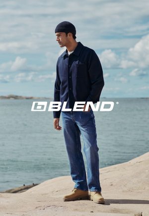 Marineblaues Button-Up-Jacket mit Brusttasche, weißes T-Shirt, blaue Jeans und beige Schuhe; das Modell trägt eine marineblaue Strickmütze und steht am Wasser.