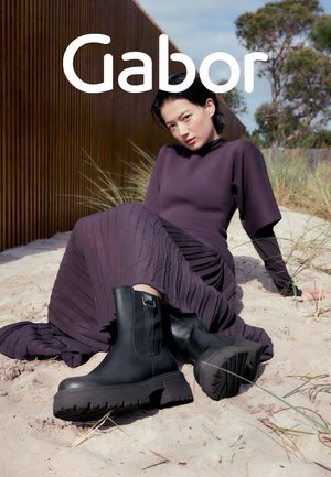 Black ankle boots with textured soles and vertical accents, worn with a long, pleated, purple dress against a sandy background.