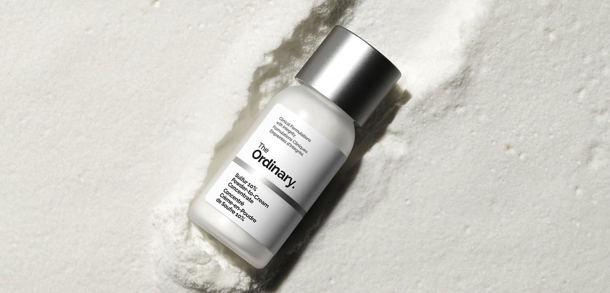 Polvere di zolfo 10% trasformata in crema, all'interno di una bottiglia bianca satinata con un tappo argentato, etichettata "The Ordinary", poggiata su una superficie bianca texturizzata.