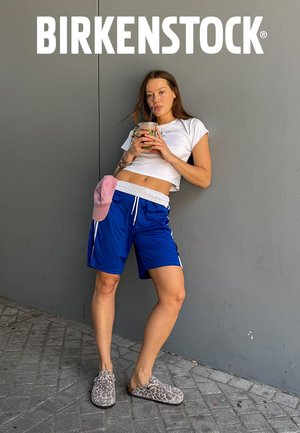 Junge Frau in blauen Shorts, weißem Croptop, rosa Hut im Bund gesteckt und Leopardenmuster-Birkenstock-Schuhen lehnt an einer grauen Wand und trinkt einen Smoothie.