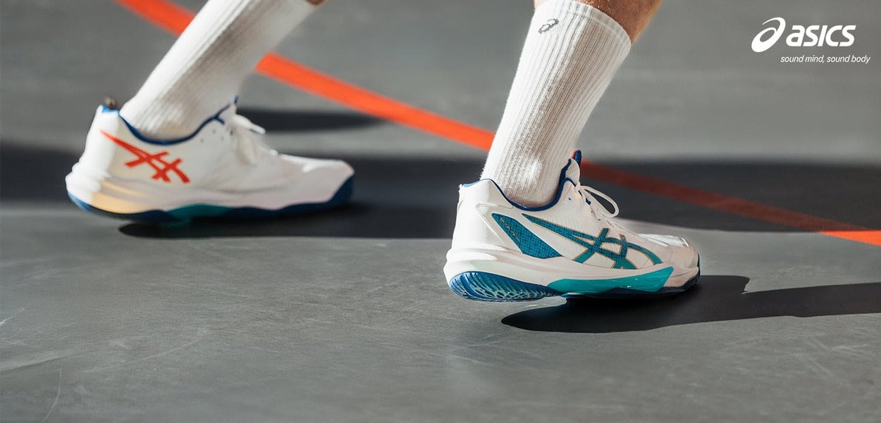 Føtter iført hvite Asics sportssko med blå og oransje detaljer på en grå innendørsbane med oransje linjer.