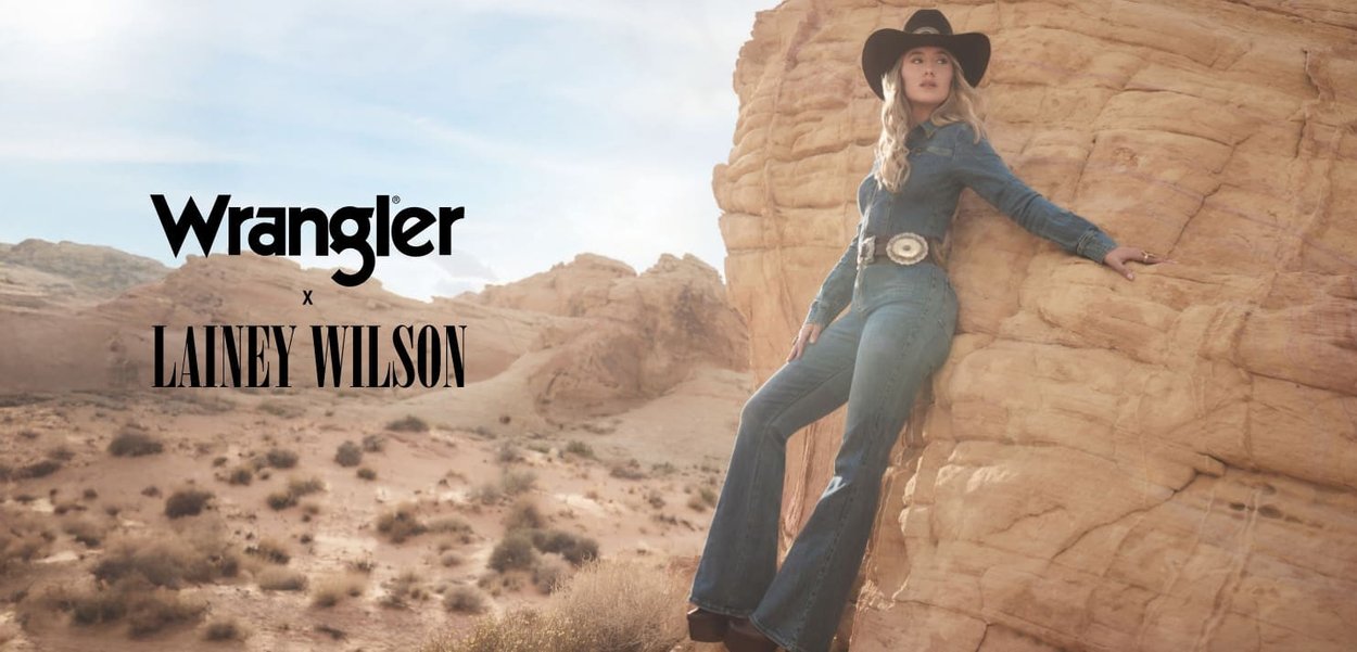 Denim jumpsuit i blått med flagrende ben, bred krage og innsnevret midje, kombinert med et stort sølvbelte og en svart cowboyhatt.