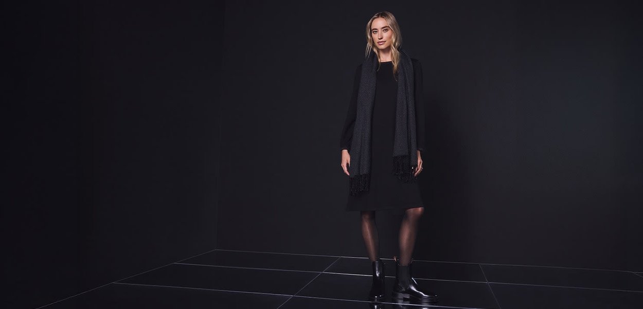 Robe noire à manches longues, associée à une écharpe foncée avec des accents de franges. Le mannequin porte des bottines. Fond sombre.