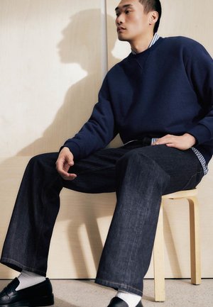 Młody mężczyzna ubrany w granatowy sweter, pasiastą koszulę, szerokie ciemne dżinsy oraz czarne lordsy, siedzący na jasnym drewnianym stołku na tle beżowej ściany.