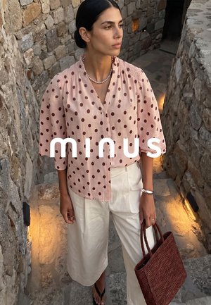 Vrouw draagt een lichtroze blouse met stippen en witte broek, houdt een bruin gevlochten handtas vast, staand op stenen treden met warm licht.