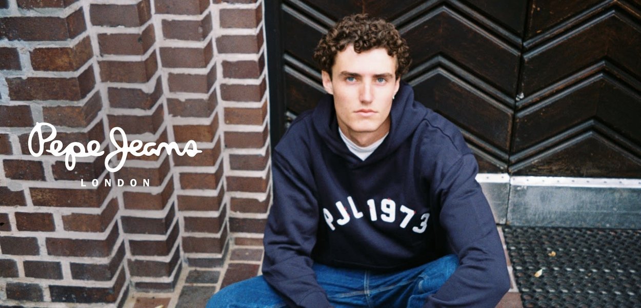 Marine hoodie met witte letters "PJL1973" gecombineerd met een blauwe spijkerbroek. Het model zit tegen een getextureerde donkere deur en bakstenen muur.