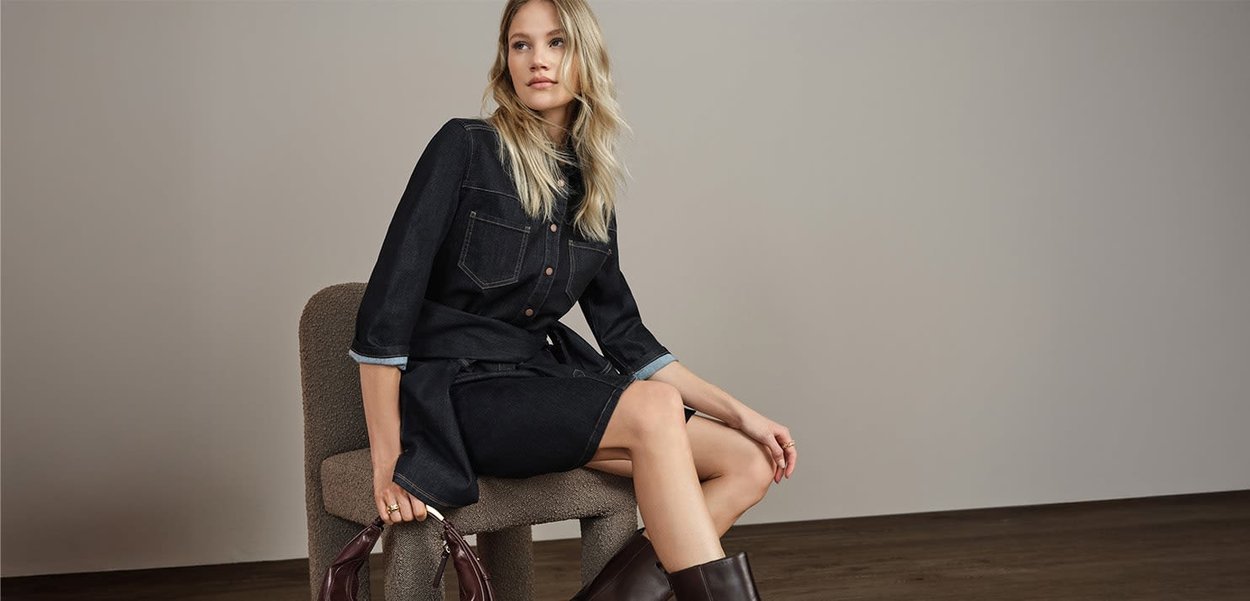Robe en denim foncé avec deux poches poitrine et une taille ceinturée, associée à des bottes marron jusqu'aux genoux et un sac à main bordeaux, assise sur une chaise claire.