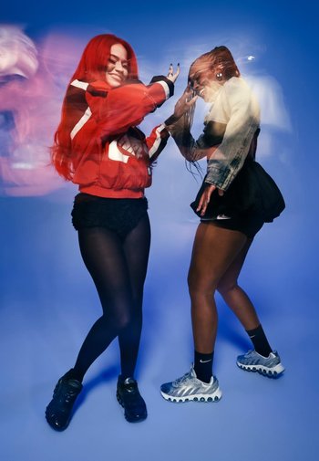 Zwei Personen posieren vor einem blauen Hintergrund. Eine trägt eine rote Jacke und schwarze Shorts; die andere ein Denim-Oberteil und einen Rock. Beide tragen Sportschuhe.
