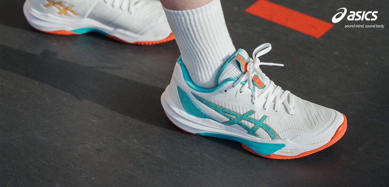 Hvite og blå ASICS sportsko med oransje såle, iført hvite ribbestrikkede sokker på et mørkt banegulv med røde markeringer.
