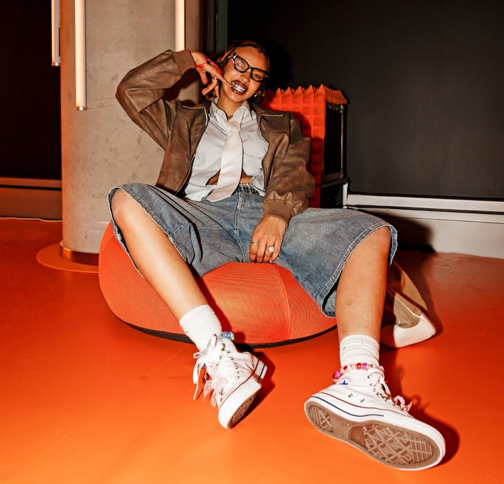 Brown leather jacket over a light blue button-up shirt and white tie, paired with denim shorts and white sneakers on an orange bean bag.