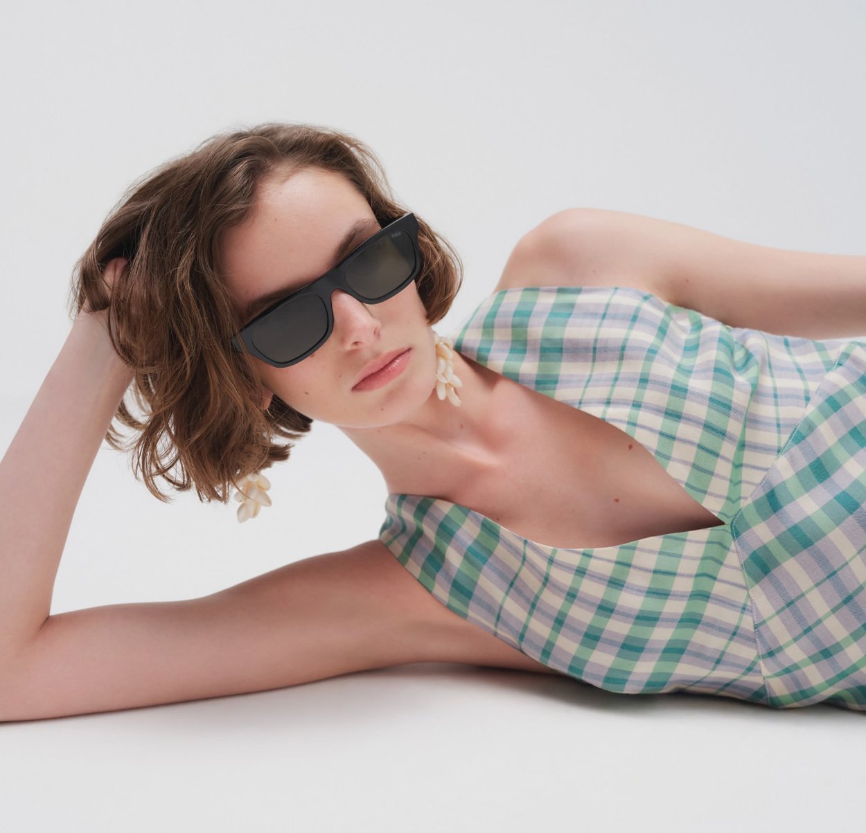 Kvinne med kort bølgete hår iført svarte solbriller, skjell-øreringer og en grønn- og hvitrutete, ermeløs topp, som lener seg tilbake på en hvit overflate.