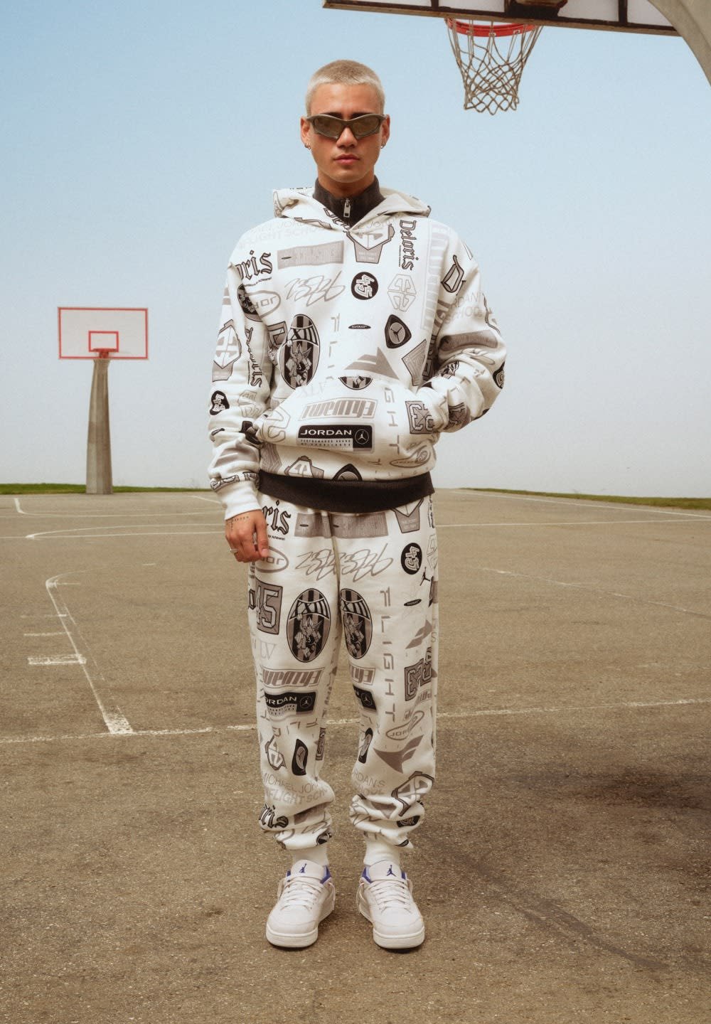 Haut à capuche blanc et pantalon avec divers logos noirs, tissu texturé et cordon de serrage. Baskets blanches avec accents bleus. Fond de terrain de basketball.