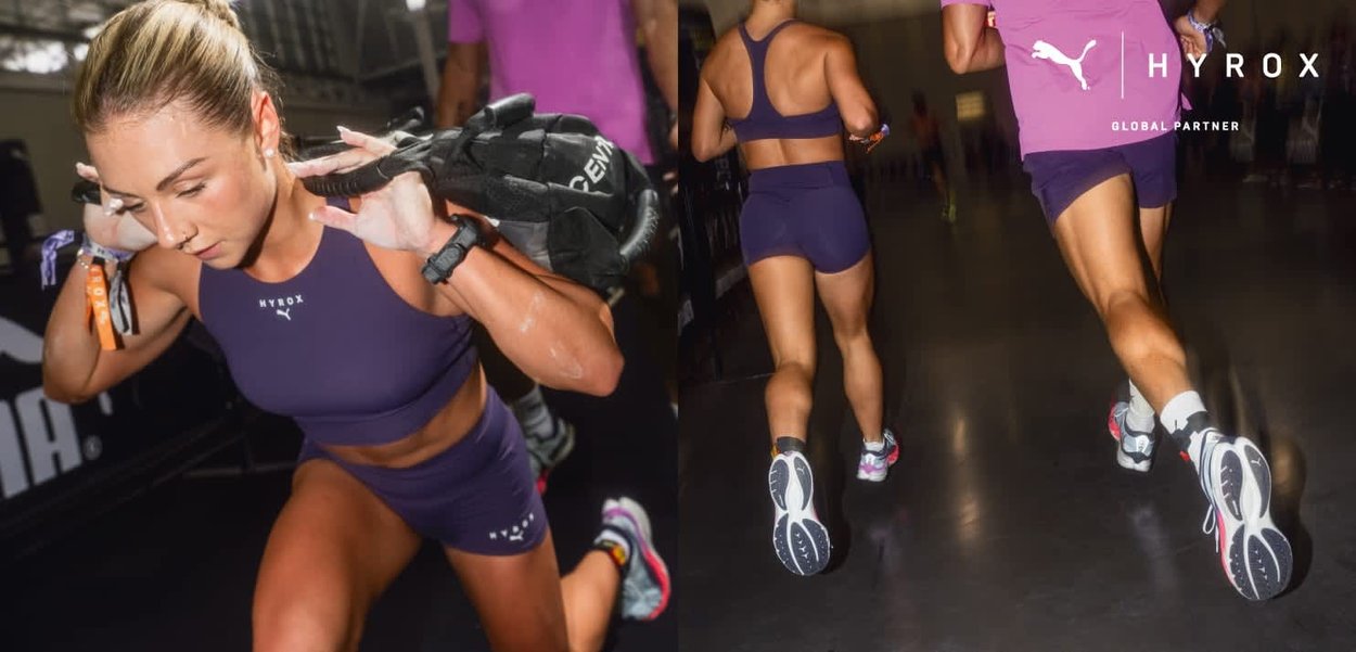 Donna in abbigliamento sportivo viola che esegue un affondo portando una sacca di sabbia, correndo accanto a un'altra atleta durante un evento di fitness indoor.