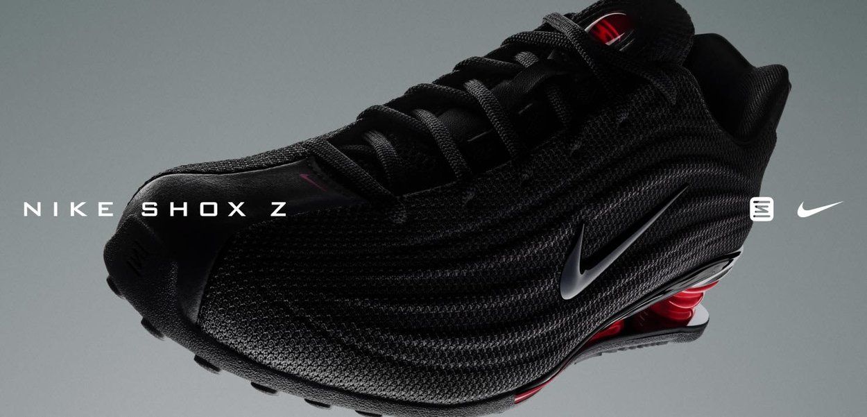 Zapatilla Nike Shox con upper de malla negra, diseño elegante con acentos en rojo, coiled de amortiguación prominente y el icónico logo plateado de Nike.