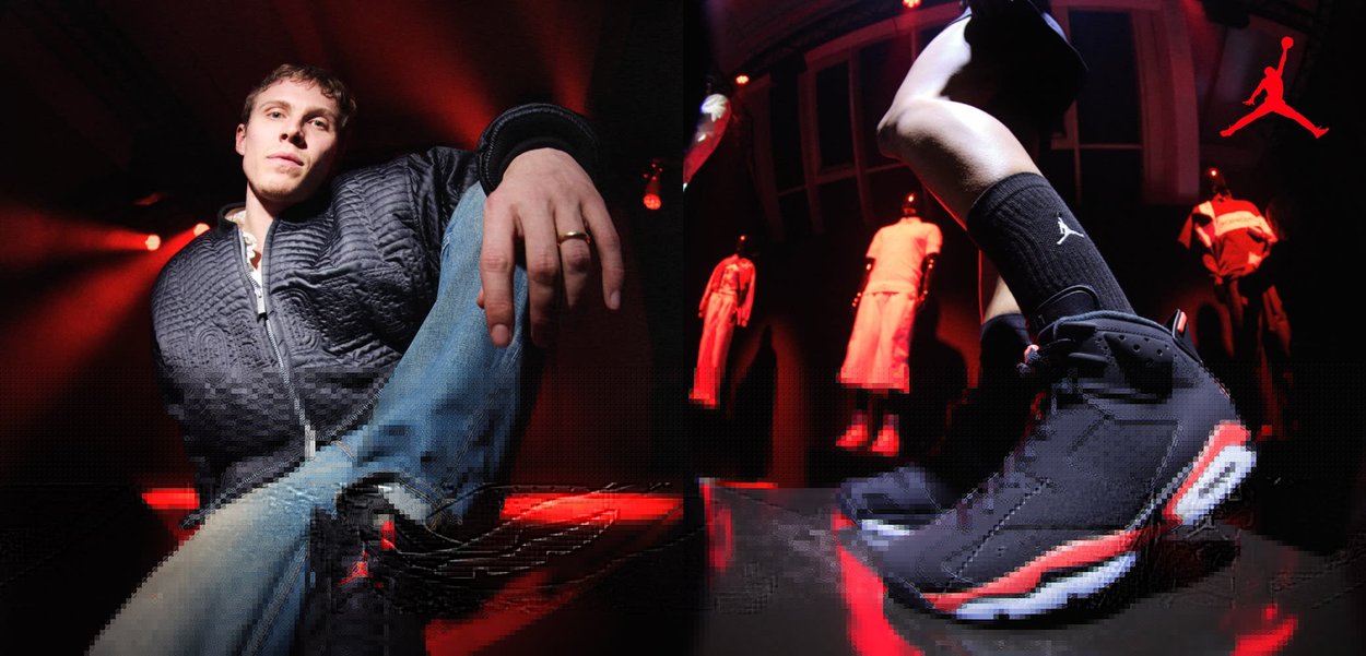 Joven vestido con chaqueta acolchada, vaqueros y zapatillas negras Air Jordan, sentado con una rodilla levantada frente a un fondo iluminado en rojo.