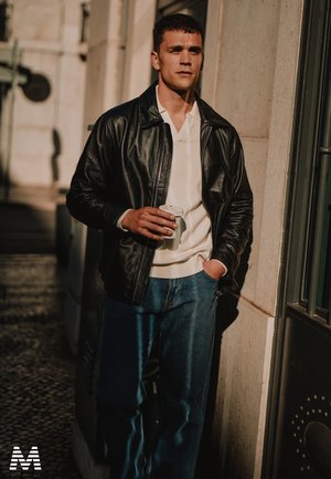 Schwarze Lederjacke über einem weißen Pullover, kombiniert mit blauen Jeans. Hält eine Kaffeetasse und steht im sanften Sonnenlicht an einer Wand.