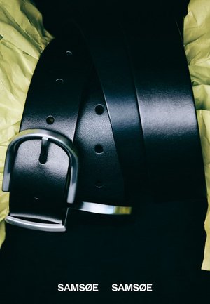 Sort læderbælte med glat tekstur og spænde i sølvmetal. Har flere huller til justering af størrelse. Vist foldet på krøllet gult papir.