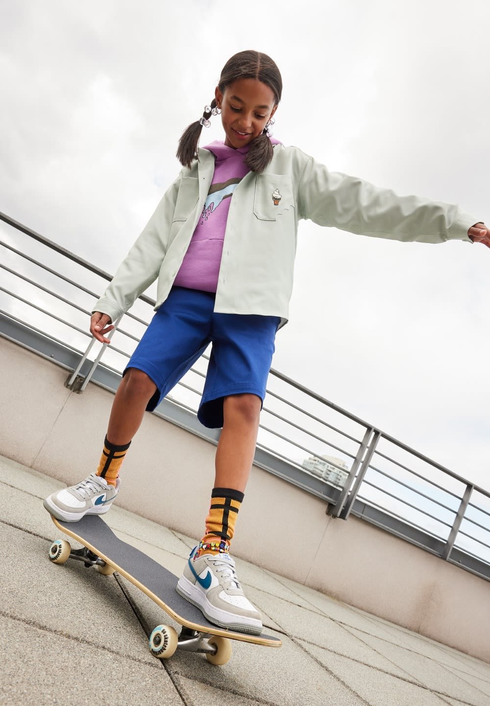 Mint green button-up shirt over purple hoodie, blue shorts, black and yellow striped socks, and white sneakers on a skateboard outdoors.