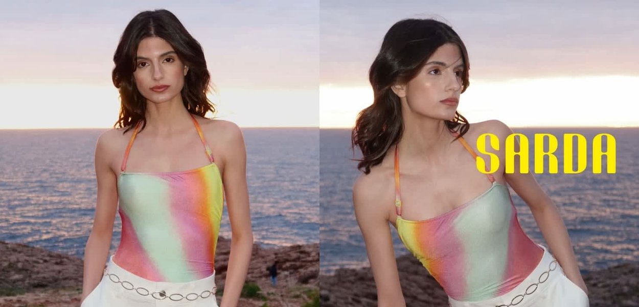 Mujer con cabello oscuro que lleva un top halter colorido y una falda blanca, de pie junto a la costa rocosa con el océano y el atardecer de fondo.