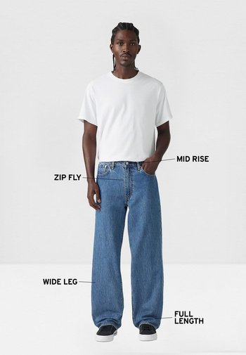 Vaqueros de mezclilla azul de pierna ancha con cintura de altura media, cierre de cremallera y diseño de longitud completa. El modelo lleva una camiseta blanca lisa y zapatillas negras.