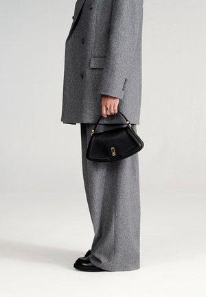 Schwarze Lederhandtasche mit strukturierter Form und goldenen Beschlägen, gehalten von einer Person in einem grauen Oversized-Blazer und Hosen.
