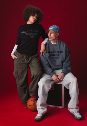 T-shirt nera dei Chicago Bulls e pantaloni cargo verdi su un modello. Felpa grigia dei Los Angeles Lakers e jeans chiari su un modello seduto. Sfondo rosso.