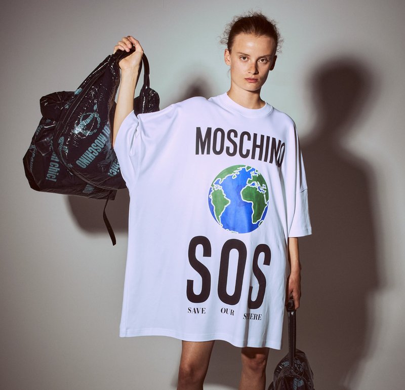 Przestronna biała koszulka z nadrukiem "MOSCHINO" i "SOS" w kolorze czarnym. Na przodzie znajduje się kolorowa grafika kuli ziemskiej. Model trzyma czarną torbę.