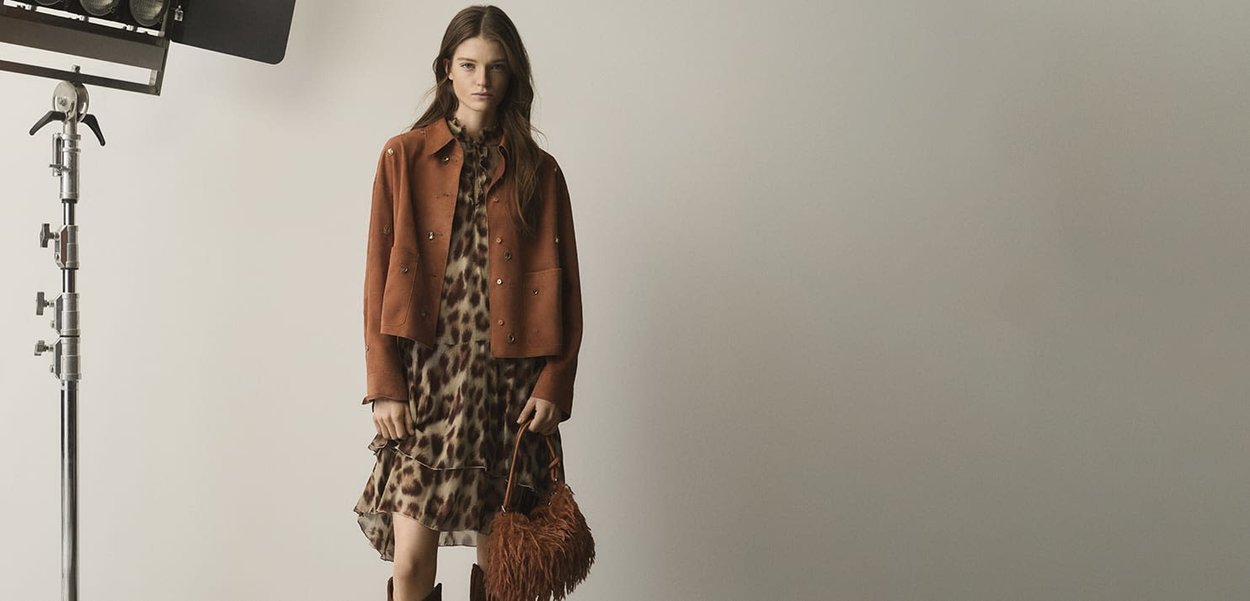 Brown suede jacket with silver buttons, leopard print dress, and a textured brown handbag. Studio lighting with a soft beige background.