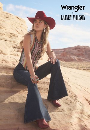 Top Wrangler x Lainey Wilson con un motivo a righe multicolori, pantaloni in denim svasati, cappello da cowboy rosso e gioielli con accenti turchesi.
