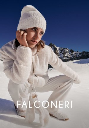 Frau mit weißer Strickmütze, Jacke und Hose hockt im Schnee, im Hintergrund verschneite Berge und klarer blauer Himmel.