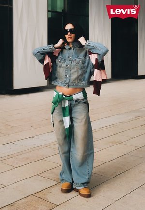 Femme portant une veste en denim oversize et un jean, avec une chemise rayée verte et blanche nouée autour de la taille, ajustant ses lunettes de soleil dans un cadre urbain.