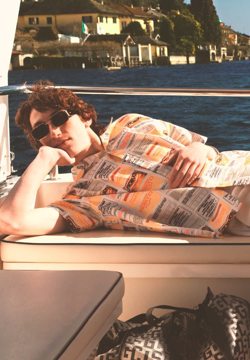 Camicia a maniche corte con un motivo colorato di etichette alimentari; il modello è sdraiato su un sedile di barca color nocciola con una borsa scura nelle vicinanze; indossa occhiali da sole.