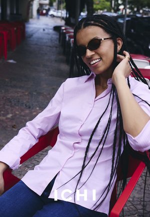 Frau mit langen Zöpfen, die Sonnenbrille trägt und ein hellviolettes Hemd mit Knöpfen trägt, sitzt draußen in einem Café auf einem roten Stuhl.