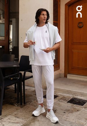 White t-shirt with logo, light gray sweater draped over shoulders, gray sweatpants, and gray sneakers. Standing on a stone surface.