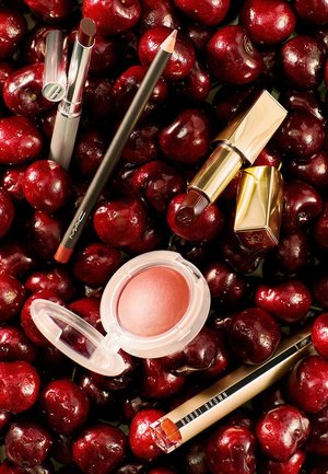 Dark red lipsticks, a lip liner, a blush compact, and an eyeliner pencil arranged on fresh red cherries.