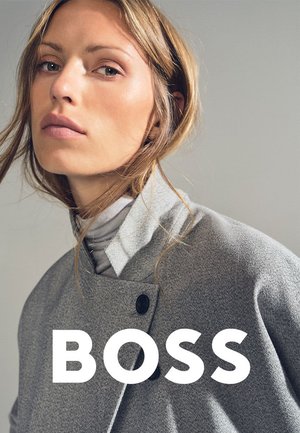 Frau mit hellbraunen Haaren, die einen grauen Mantel und einen Rollkragenpullover trägt, leicht zur Seite blickend vor einem schlichten Hintergrund, mit dem Schriftzug "BOSS".