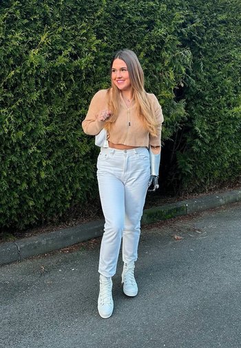 Beige kortärmad tröja med ribbad textur, vita högmidjade jeans och vita högtoppsskor. Synlig detaljer av protesarm. Grön häck i bakgrunden.