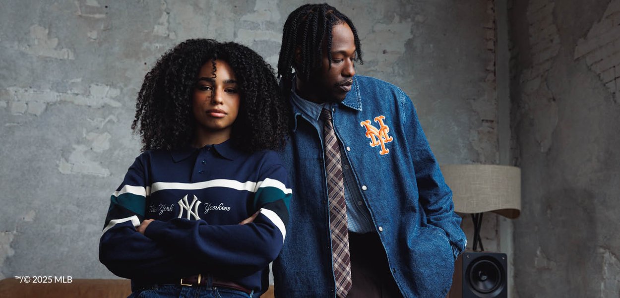 Kvinde iført en New York Yankees trøje med armene over kors står ved siden af en mand i en denimjakke fra New York Mets, indendørs mod en tekstureret væg.