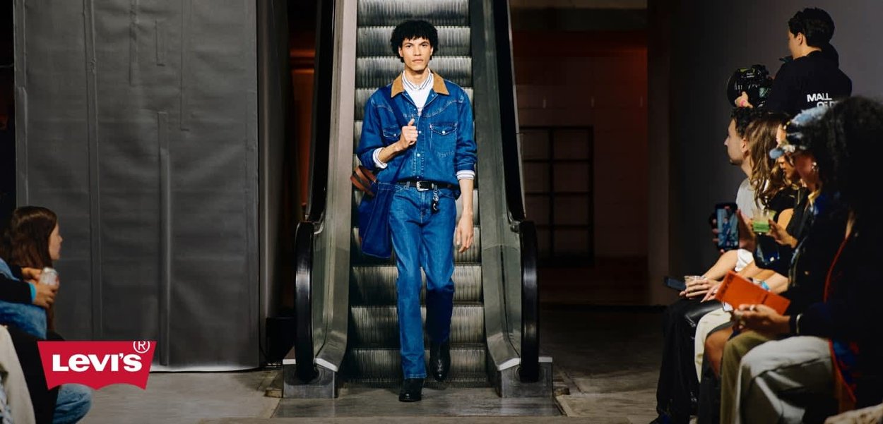 Model in a blue denim jacket with a tan collar, blue jeans, and black shoes, walking down an escalator during a fashion show.