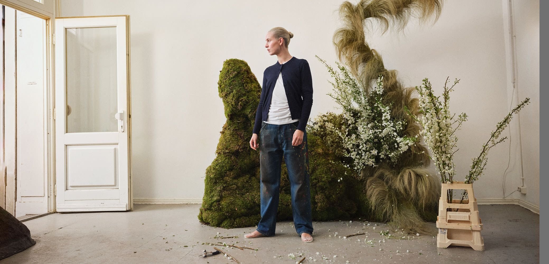 Model staat in een kamer naast een gestructureerde groene mos en hoge, veerachtige planten, met witte bloemen en een lichthouten kruk.