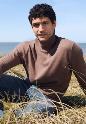 Junger Mann mit dunklem, lockigem Haar, der einen braunen Rollkragenpullover und blaue Jeans trägt, sitzt im hohen Gras am Meer unter einem klaren Himmel.