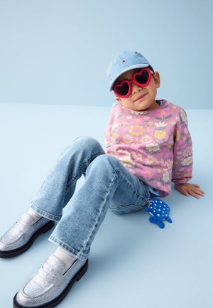 Bambino che indossa occhiali da sole a forma di cuore rossi, maglione floreale rosa, cappellino blu, jeans e scarpe argento, seduto su un pavimento azzurro chiaro con un giocattolo blu a pois.