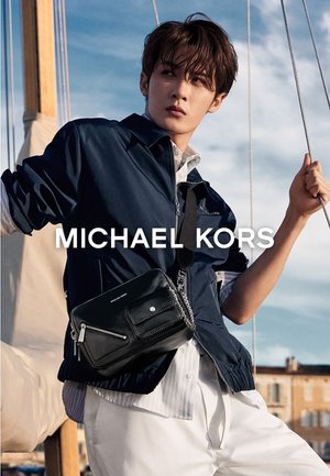 Junger Mann auf einem Boot, der eine marineblaue Jacke, weiße Hosen und eine schwarze Michael Kors Umhängetasche trägt und ein Segelseil gegen den blauen Himmel hält.