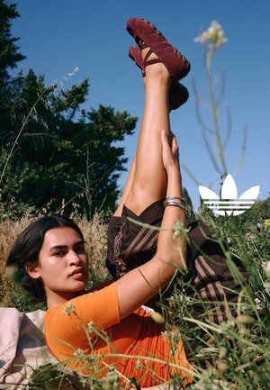 Jeune femme allongée dans l'herbe à l'extérieur avec les jambes levées, portant un haut orange, un pantalon marron et des chaussures bordeaux sur fond de ciel bleu.