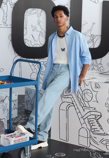 Hellblau gestreiftes Hemd über einem weißen T-Shirt mit Schmetterlingsdesign, kombiniert mit hellblauen Jeans und schwarzen Sneakers, lehnt an einem Wagen.
