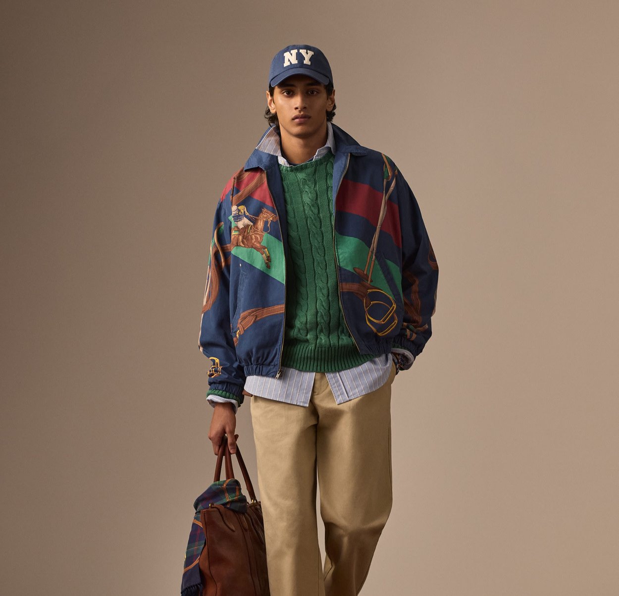 Veste bomber colorée avec des imprimés équestres, pull en mailles torsadées vert, chemise bleu à boutons, pantalon beige et sac fourre-tout marron.