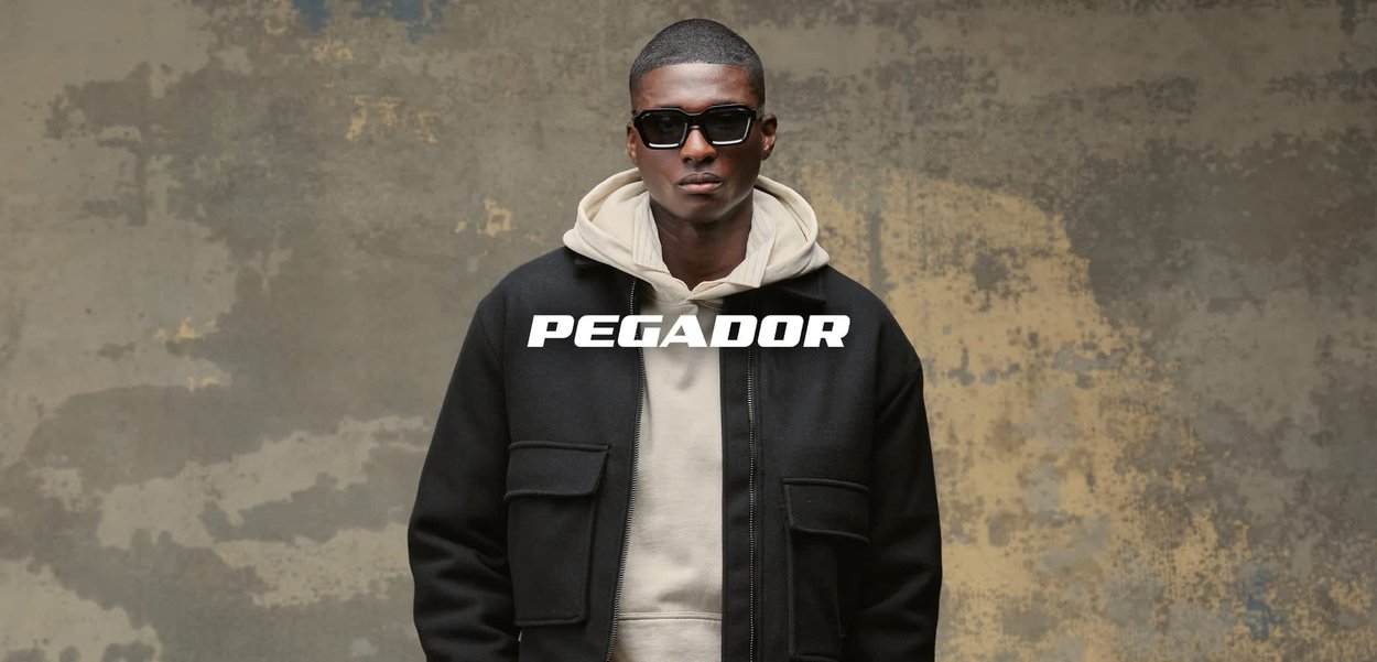 Giacca nera con cerniera e due tasche frontali, indossata sopra una felpa con cappuccio beige chiaro. L'outfit comprende pantaloni beige chiaro. Lo sfondo è strutturato.