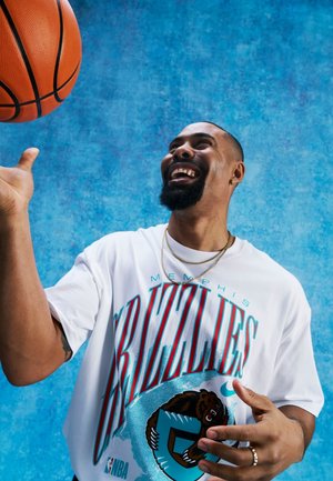 Hombre haciendo girar un balón de baloncesto en el dedo, vistiendo una camiseta blanca de Memphis Grizzlies frente a un fondo azul texturizado.