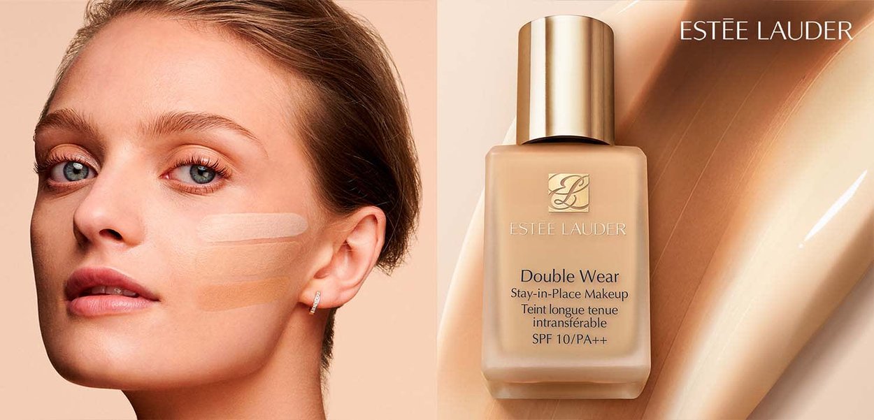 Donna con occhi azzurri e pelle chiara mostra tre campioni di fondotinta sulla guancia accanto a una bottiglia di fondotinta Estee Lauder Double Wear.
